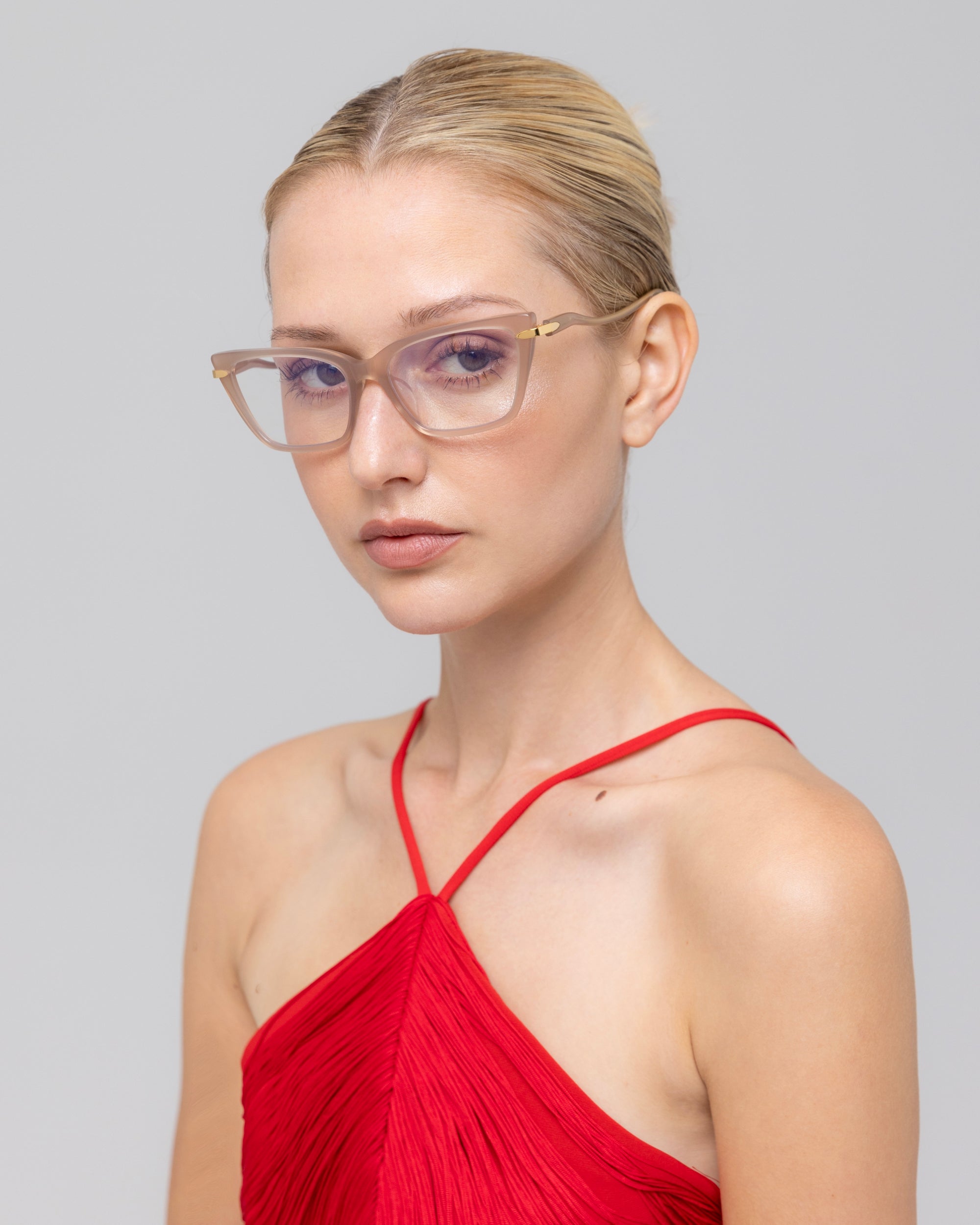 A woman with blonde hair in a ponytail wears For Art's Sake® Motion I clear-framed glasses with acetate temples and a red halter-neck top, gazing to the side against a plain light gray background.