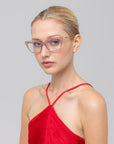 A woman with blonde hair in a ponytail wears For Art's Sake® Motion I clear-framed glasses with acetate temples and a red halter-neck top, gazing to the side against a plain light gray background.