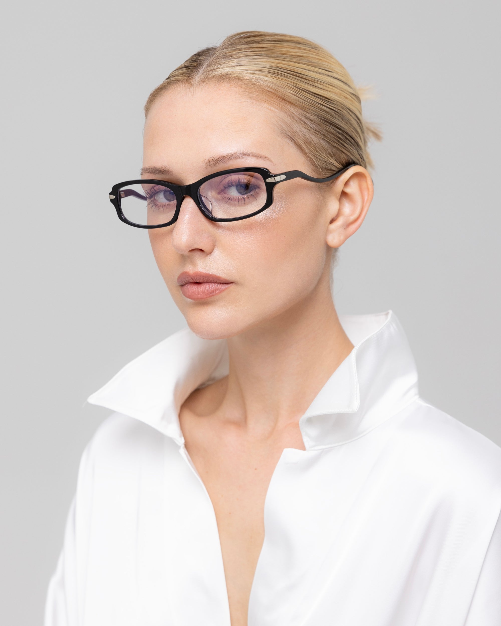 A blonde woman with a neat bun wears For Art's Sake® Motion II rectangular sunglasses with curved acetate temples and a white collared shirt, looking confidently at the camera against a plain light gray background.