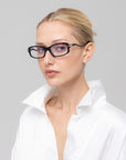 A blonde woman with a neat bun wears For Art's Sake® Motion II rectangular sunglasses with curved acetate temples and a white collared shirt, looking confidently at the camera against a plain light gray background.