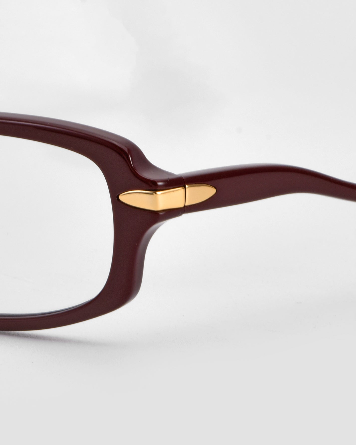 Close-up of For Art&#39;s Sake® Motion II eyeglasses featuring curved acetate temples and a small gold oval metal accent by the hinge. The dark brown or burgundy frame contrasts with a plain, light backdrop.