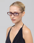 A woman with blonde hair in a neat bun wears For Art's Sake® Motion II maroon rectangular glasses with curved acetate temples and a black halter top, looking slightly to the side against a plain light gray background.
