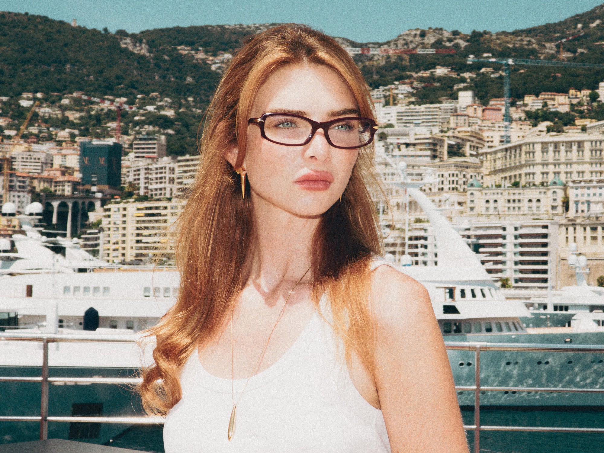 A woman with long, wavy blonde hair wears For Art's Sake® Motion II glasses with acetate temples and a white tank top, standing before yachts and buildings on a sunny day, hills in the background.