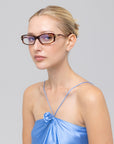 A woman with blonde hair in a bun wears For Art’s Sake® Motion II boxy rectangular sunglasses with metal accents and a light blue satin spaghetti-strap top, gazing neutrally at the camera against a plain gray background.