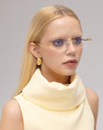 A woman with long blonde hair showcases For Art's Sake® Ida lightweight rimless glasses with gold accents, styled with gold geometric earrings and a sleeveless high-collar pale yellow top against a neutral background.