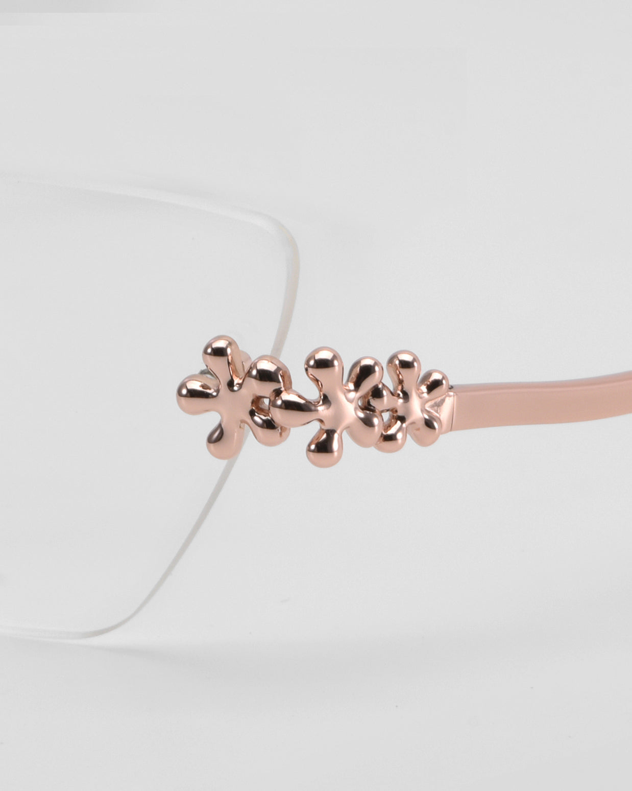Close-up of For Art's Sake® Ida eyeglasses, featuring a rose gold metal frame with three flower-shaped accents on the temple. Lightweight and clear-lensed, they display a subtle floral design against a light gray background.