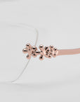 Close-up of For Art's Sake® Ida eyeglasses, featuring a rose gold metal frame with three flower-shaped accents on the temple. Lightweight and clear-lensed, they display a subtle floral design against a light gray background.