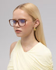 A young woman with long blonde hair poses against a light gray background wearing For Art's Sake® Claudia sunglasses—Georgia O’Keeffe inspired acetate and metal frames with sculpted temples—paired with gold earrings and a pale yellow one-shoulder top.