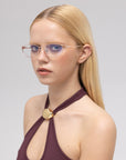 A young woman with straight blonde hair models the Anita geometric metal eyewear by For Art's Sake®, paired with a sleeveless maroon halter top featuring a gold circle detail, against a neutral gray background.