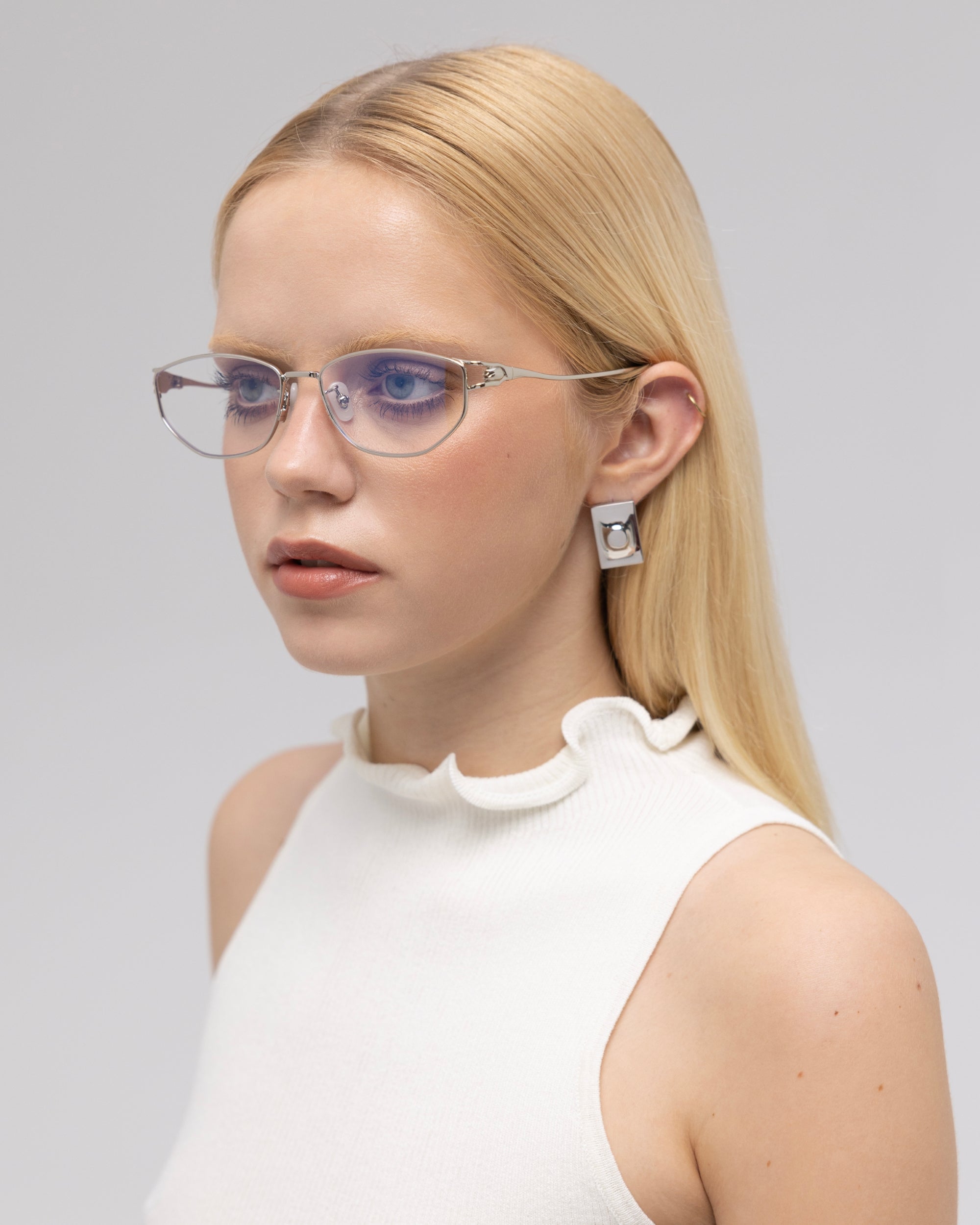 A young woman with long blonde hair wears For Art's Sake® Anita geometric metal eyeglasses, a sleeveless white top with a ruffled neckline, and geometric earrings, gazing slightly to the side against a plain light background.