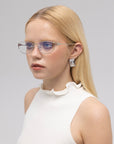 A young woman with long blonde hair wears For Art's Sake® Anita geometric metal eyeglasses, a sleeveless white top with a ruffled neckline, and geometric earrings, gazing slightly to the side against a plain light background.