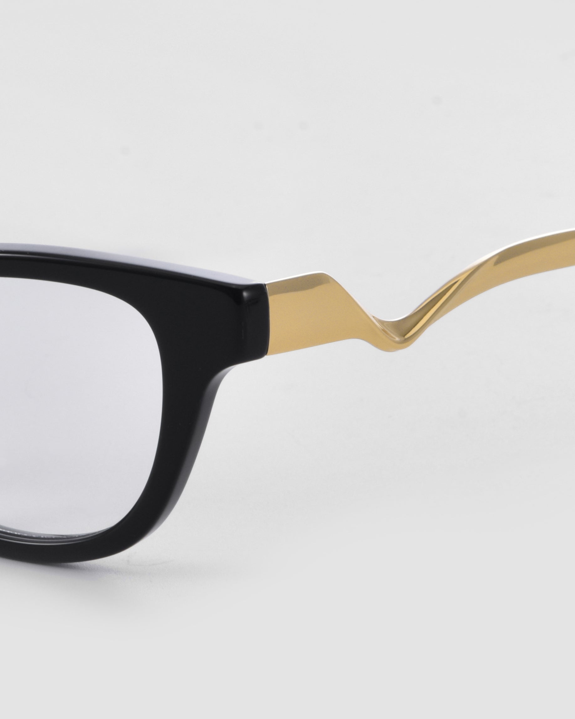 Close-up of For Art's Sake® Poppy optical frames: black square shape and gold sculptural zigzag metal temple arms. Set against a plain light gray background.