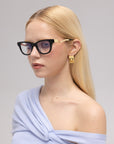 A woman with long blonde hair wears For Art's Sake® Poppy bold temple glasses, gold geometric earrings, and a light blue off-the-shoulder top, gazing slightly to the side against a plain background.