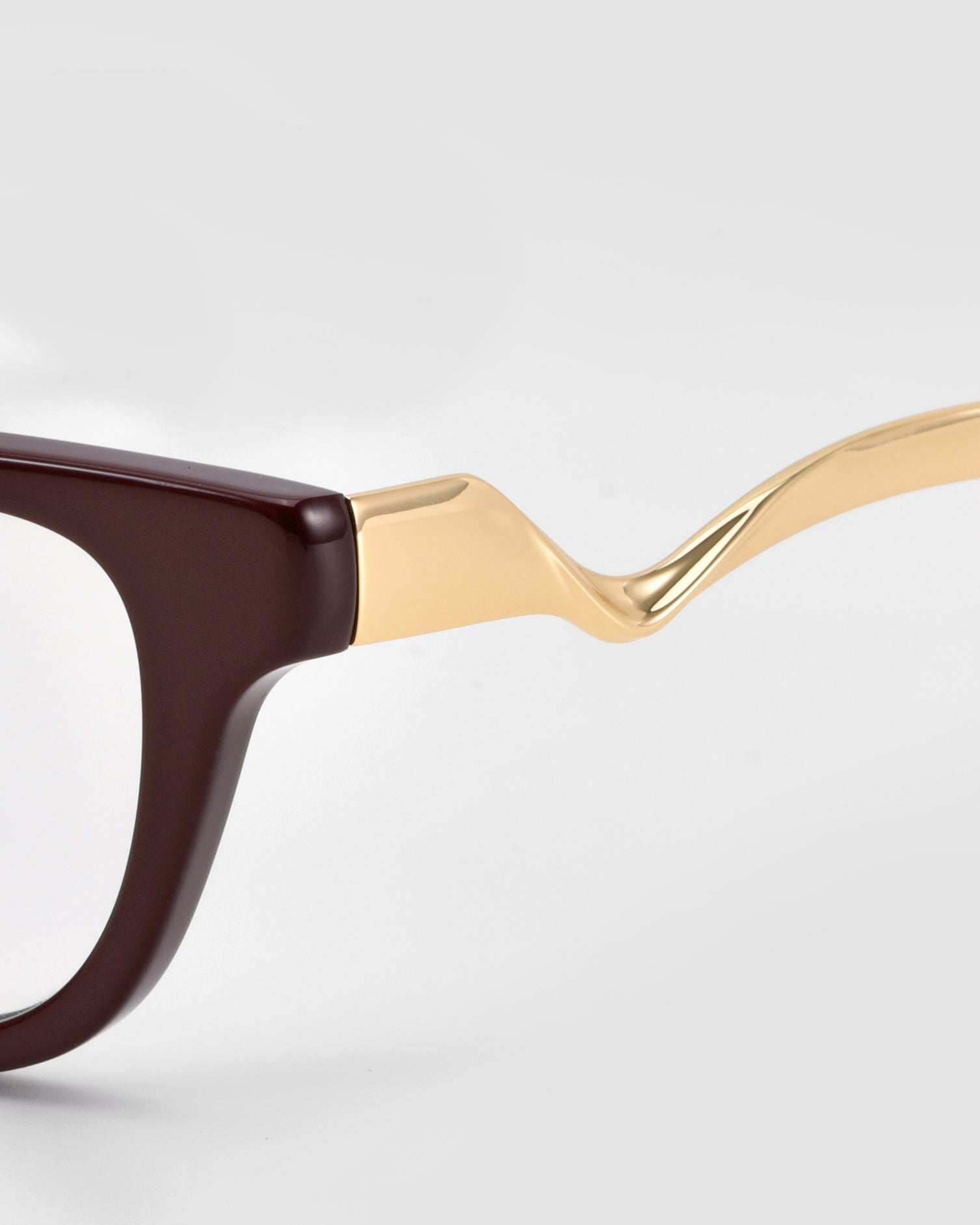 Close-up of For Art's Sake® Poppy eyeglasses featuring a dark brown front and a glossy, wavy gold sculptural metal temple, set against a light grey background.