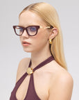 A woman with long blond hair wears For Art's Sake® Poppy statement eyewear—bold dark frames with striking temples—paired with a sleeveless maroon top, gold geometric earrings, and poses against a plain light gray background.