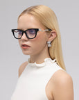 A young woman with long blonde hair wears For Art's Sake® Poppy bold temple eyeglasses, a sleeveless white top with a ruffled collar, and rectangular silver earrings, posing against a plain light gray background.