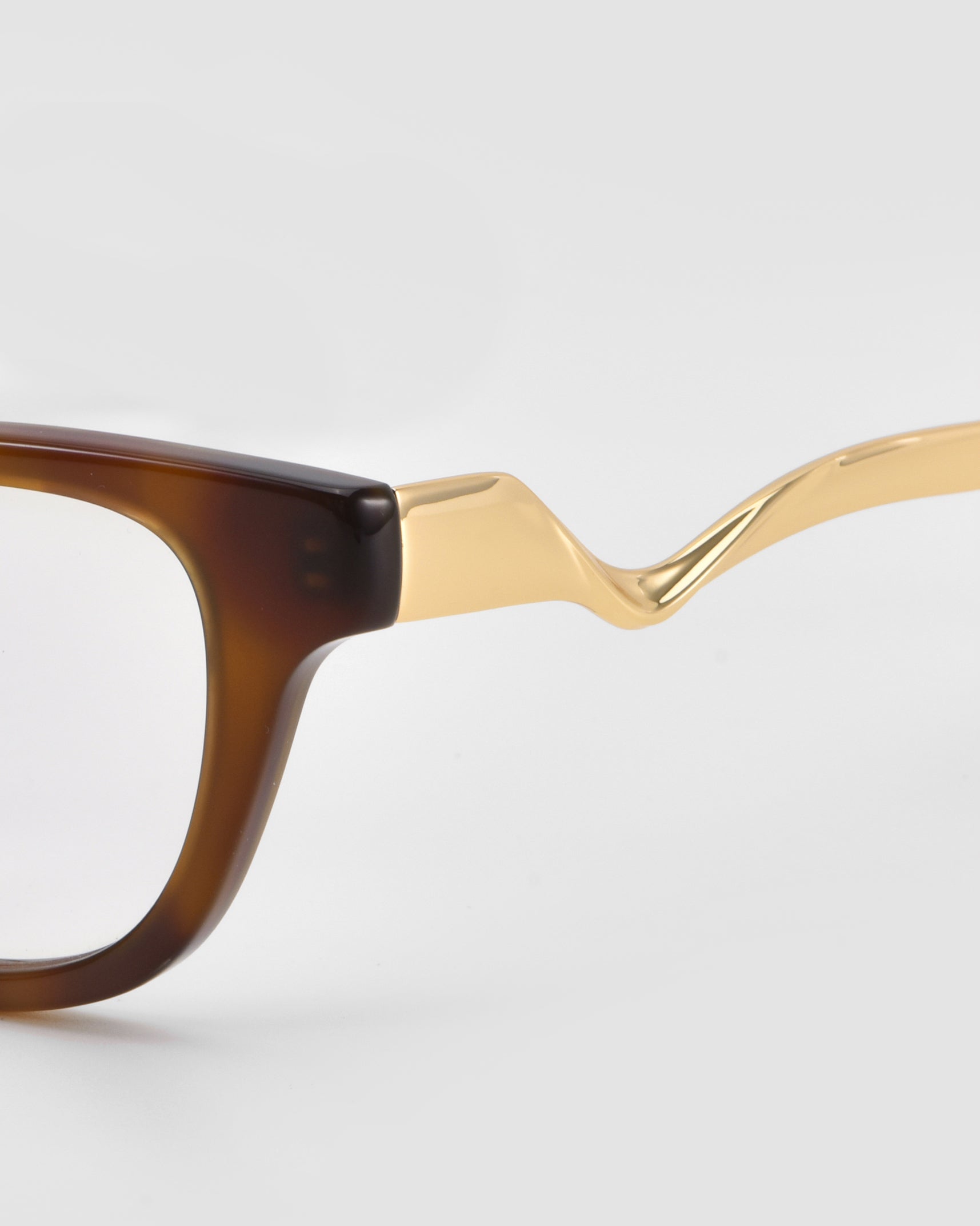 Close-up of For Art's Sake® Poppy eyeglasses with a brown rectangular frame and bold temples, featuring twisted gold sculptural metal temple arms against a light background.