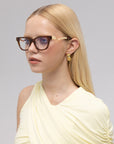 A woman with straight blonde hair wears Poppy glasses by For Art's Sake® in brown and gold, gold hoop earrings, and a pale yellow one-shoulder top. She looks slightly to the side against a light gray background.