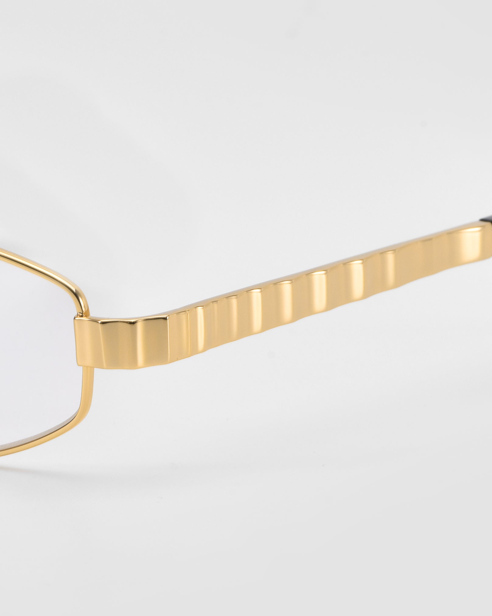 Close-up of the gold, textured arm of the Iris geometric rectangular eyewear by For Art's Sake®, set against a plain light gray background.