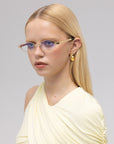 A woman with straight blonde hair wears For Art's Sake® Iris gold-rimmed blue-tinted glasses, gold hoop earrings, and a light yellow one-shoulder top against a plain light gray background.