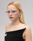 A woman with straight blonde hair models For Art's Sake® Iris eyewear—narrow, blue-tinted geometric opticals—paired with a black one-shoulder dress, a brooch, and rectangular earring against a plain light background.