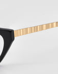 Close-up of For Art's Sake® Lavender eyewear: glossy black sculptural optical frames featuring a textured metallic gold temple arm, set against a plain light gray background.