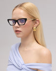 A woman with long blonde hair wears For Art’s Sake® Lavender black cat-eye acetate glasses, gold geometric earrings, and a light blue off-shoulder top, posing against a plain light background.