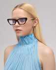 A woman with straight blonde hair wears For Art's Sake® Lavender black cat-eye acetate glasses, silver triangular earrings, and a sleeveless light blue high-neck top, posing against a plain light gray background.