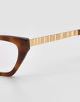 Close-up of For Art's Sake® Lavender eyeglasses featuring a brown rectangular frame and gold ribbed temple arms, set against a plain light gray background—an elegant statement of quiet luxury.