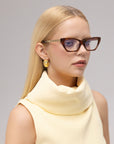 A woman with straight blonde hair wears For Art's Sake® Lavender sculptural cat-eye acetate glasses and gazes to the side, set against a neutral background.