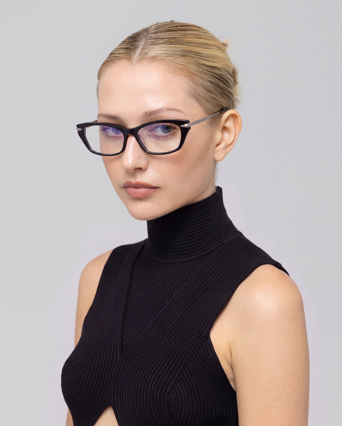 A woman with blonde hair in a sleek bun wears For Art’s Sake® Suri I black-rimmed optical frames with metal temples and a sleeveless black turtleneck, set against a plain light background.