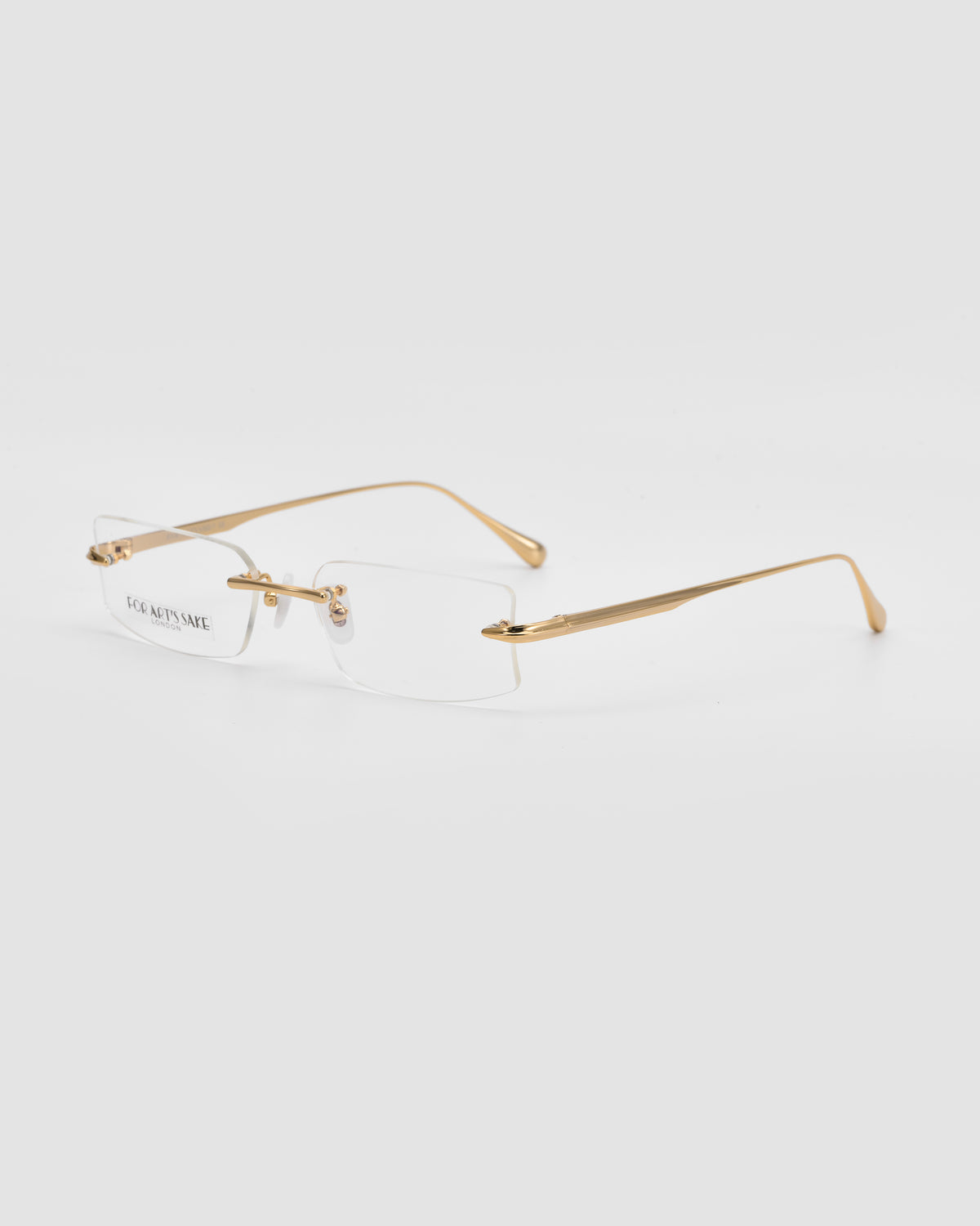 Suri II by For Art’s Sake®: Rimless glasses with rectangular clear lenses, slim gold metal temples and bridge, plus jade nose pads, shown on a plain light gray background.