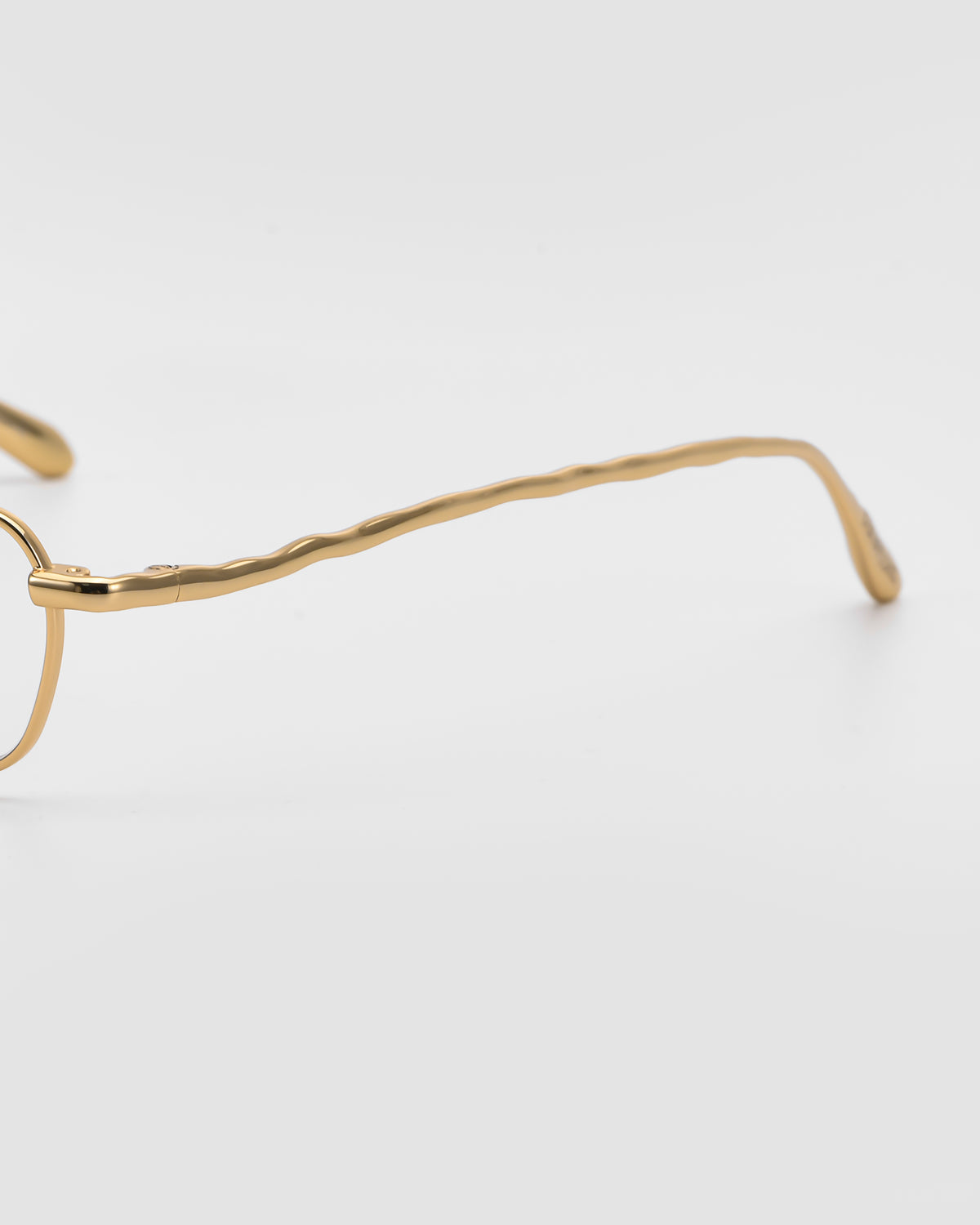 A close-up side view of For Art’s Sake® Suree I gold metal glasses with wavy, textured temples and a subtle rectangular silhouette on a white background.