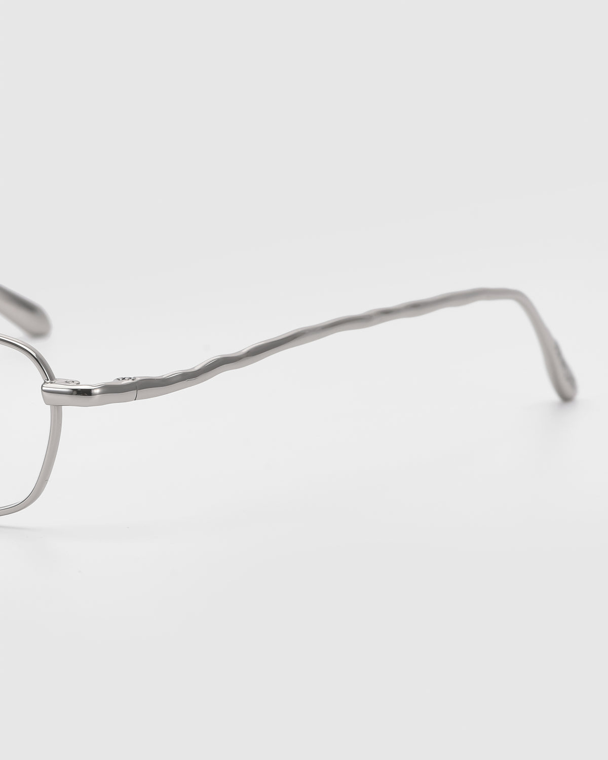 Close-up side view of Suree I by For Art’s Sake®—metal glasses with a thin, wavy-patterned temple arm and rectangular silhouette, shown on a plain white background.