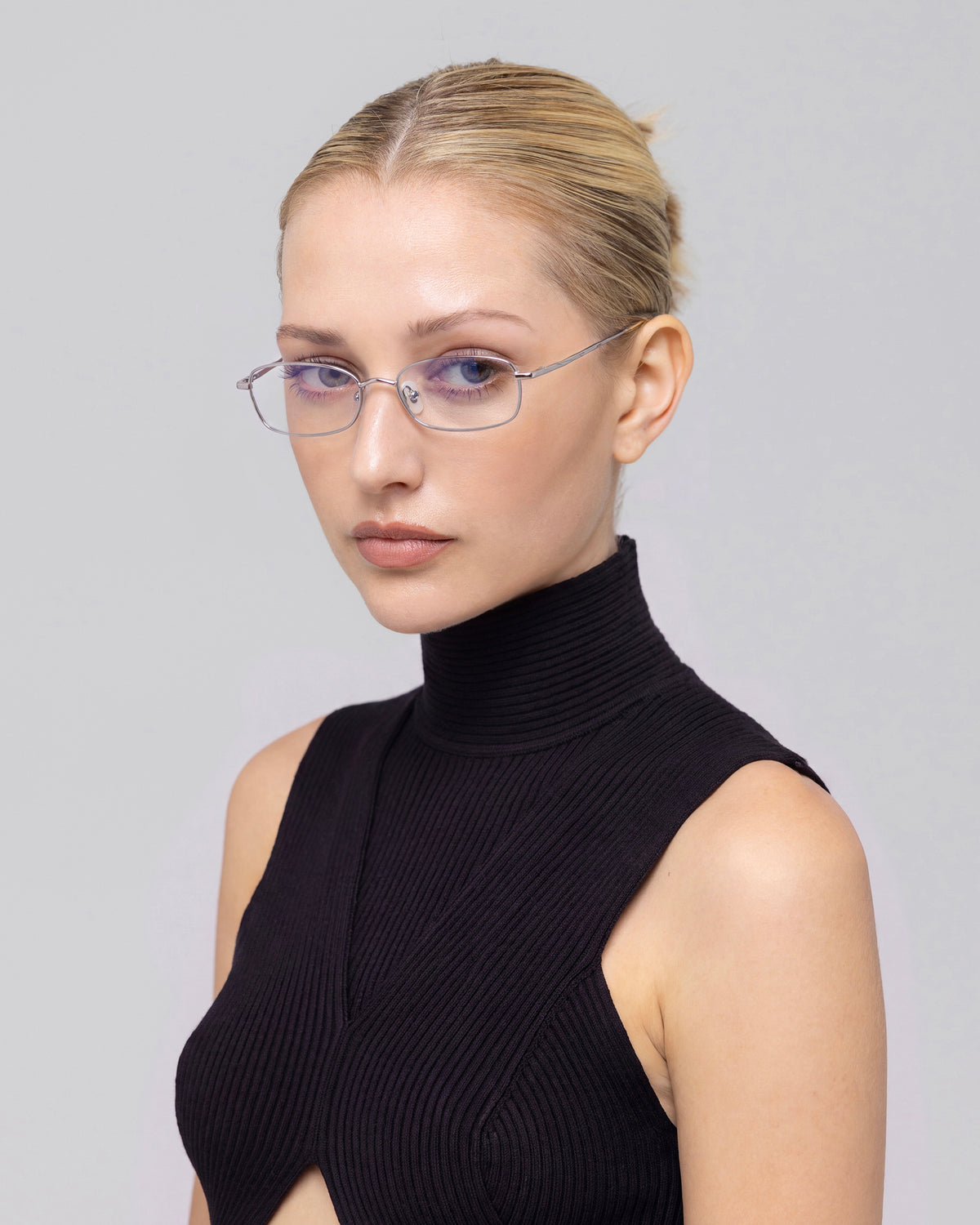 A woman with blond hair in a sleek bun wears For Art’s Sake® Suree I rectangular optical frames and a sleeveless black turtleneck, gazing to the side against a plain light background.