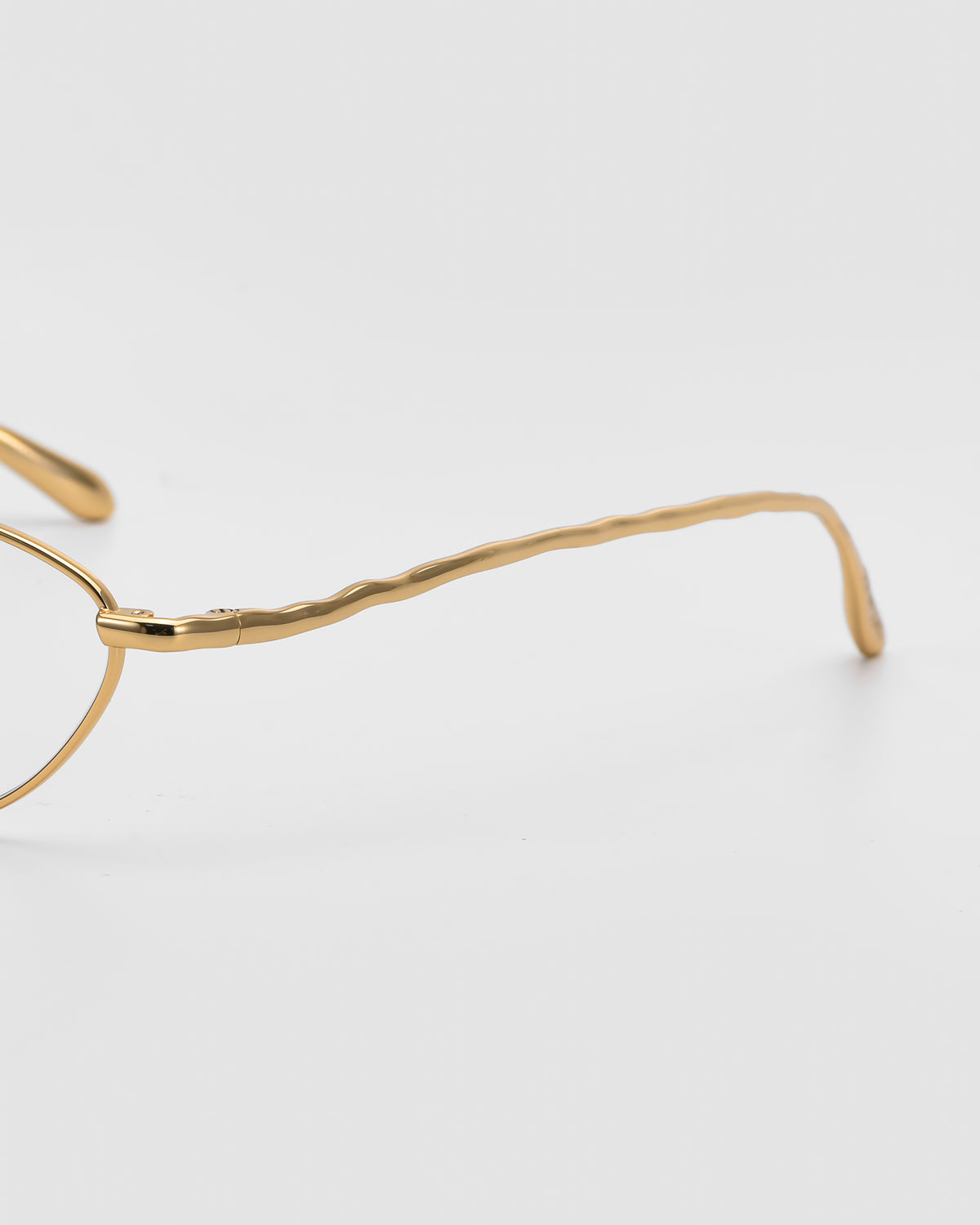 Close-up of For Art’s Sake® Suree II gold metal frames with a wavy, textured temple arm on a white background. Only a section of these elegant optical frames and the arm are shown.
