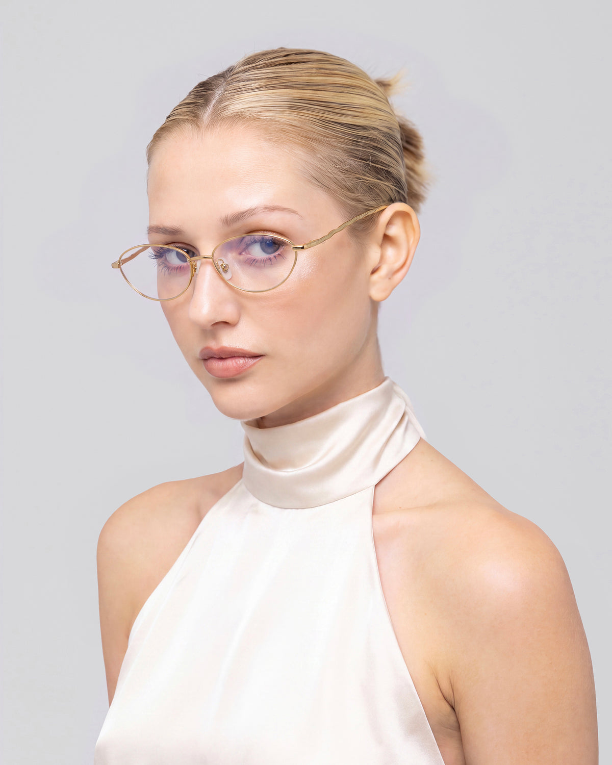 A woman with blond hair in a low bun wears For Art’s Sake® Suree II optical frames and a sleeveless, high-neck satin top, gazing at the camera against a plain light gray background.