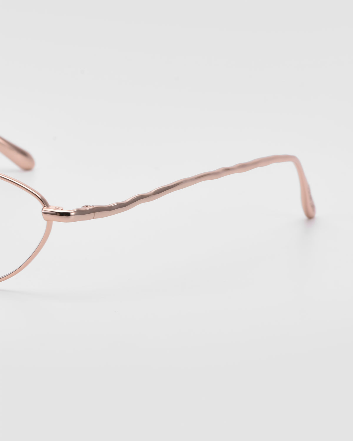 Close-up of the side of For Art’s Sake® Suree II thin rose gold optical frames, featuring a wavy pattern along the arm, set against a plain white background.