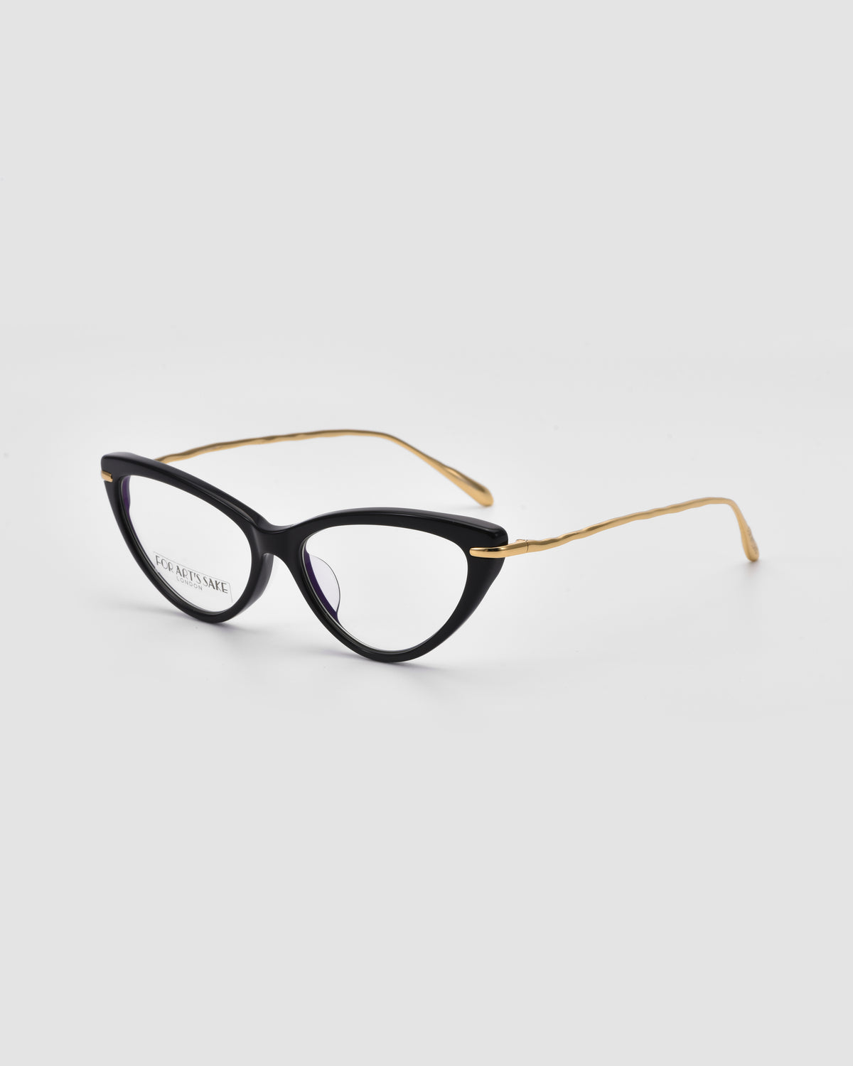Solange by For Art’s Sake® features cat-eye opticals with black frames and sculpted metal temples, resting on a light gray surface. The left lens bears text, giving these jewellery-inspired glasses a modern edge.