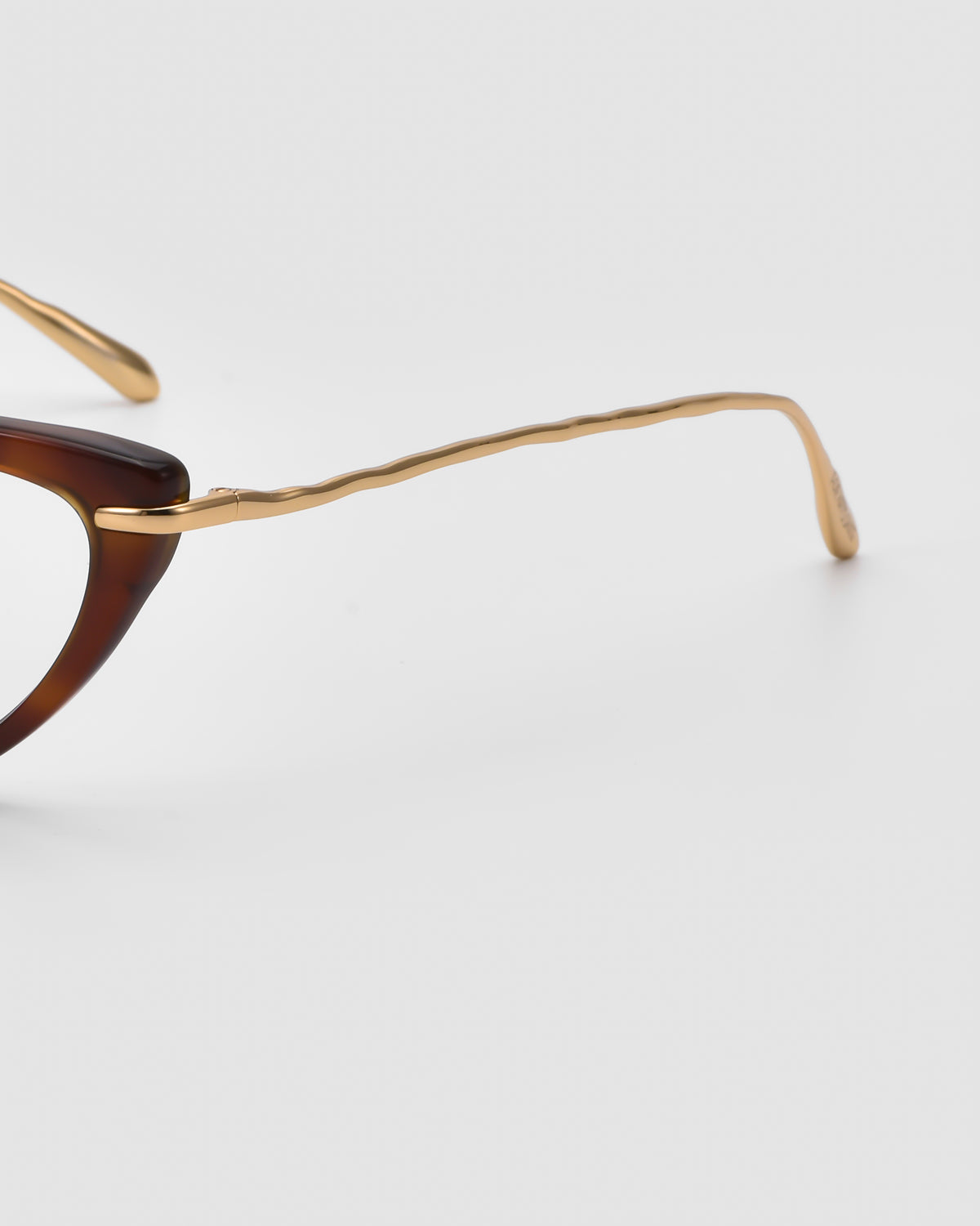 Close-up of For Art’s Sake® Solange glasses featuring a jewellery-inspired brown frame and thin, gold, sculpted metal temples, set against a plain light background.