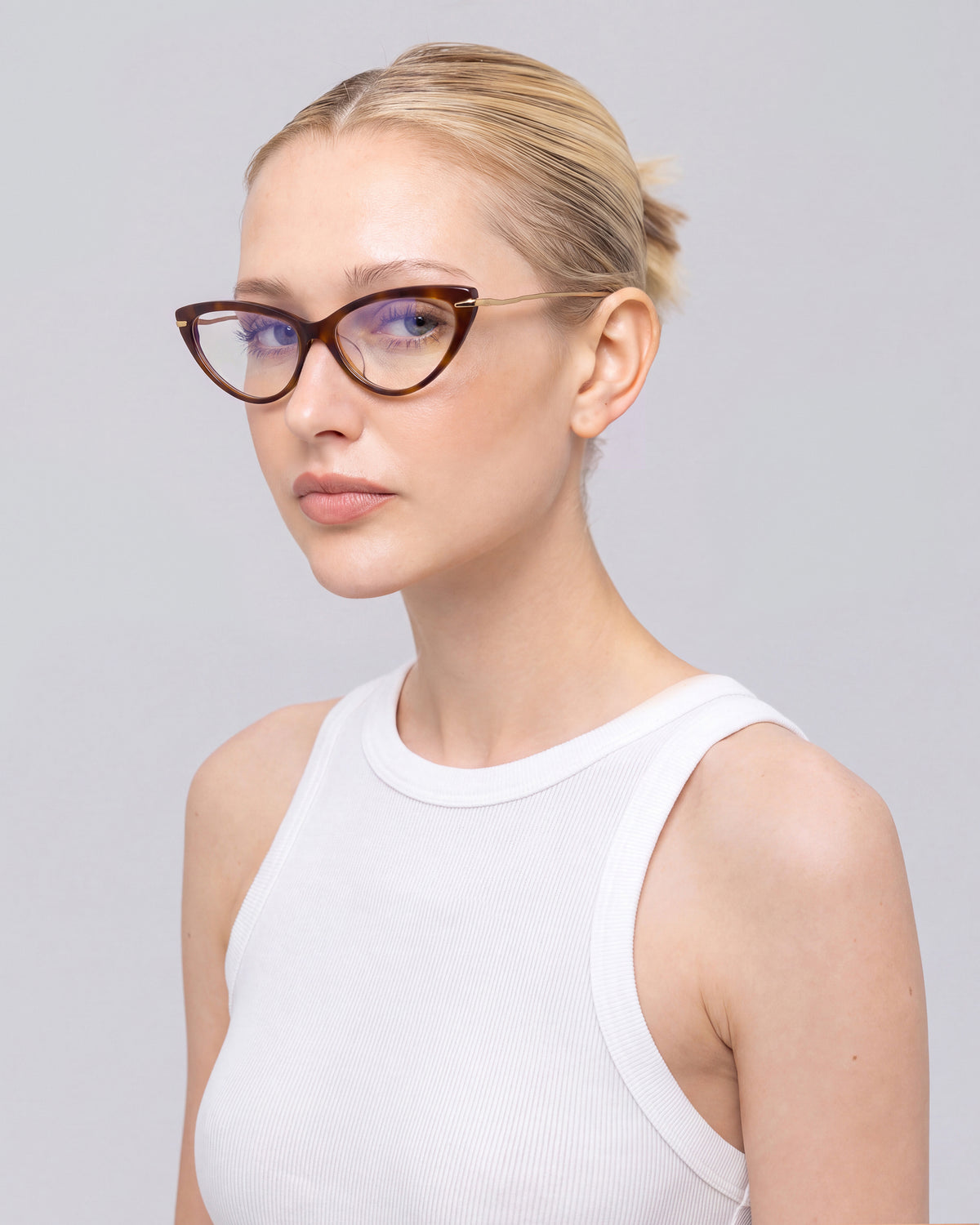 A blonde woman with her hair in a bun wears For Art’s Sake® Solange cat-eye opticals with sculpted metal temples and a sleeveless white top, gazing to the side against a plain light gray background.