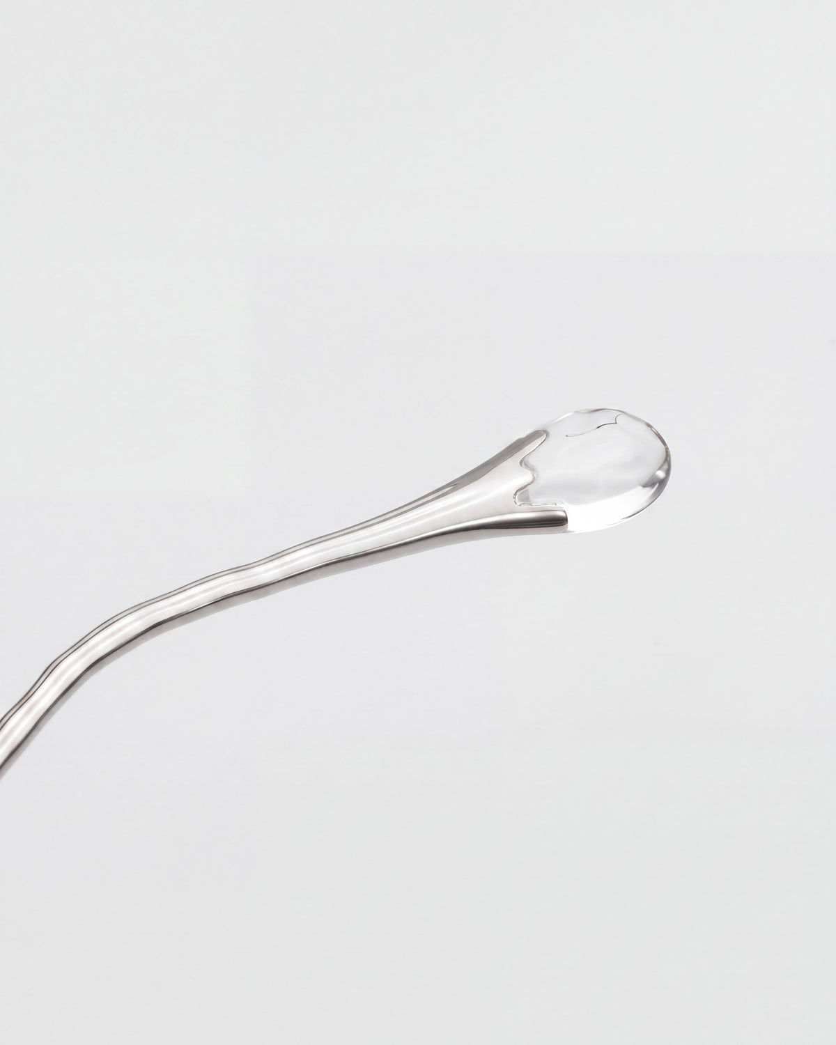 A close-up of Amara I by For Art’s Sake® shows a clear droplet on a shiny, curved metal utensil, resembling a translucent jewel against a plain white background.