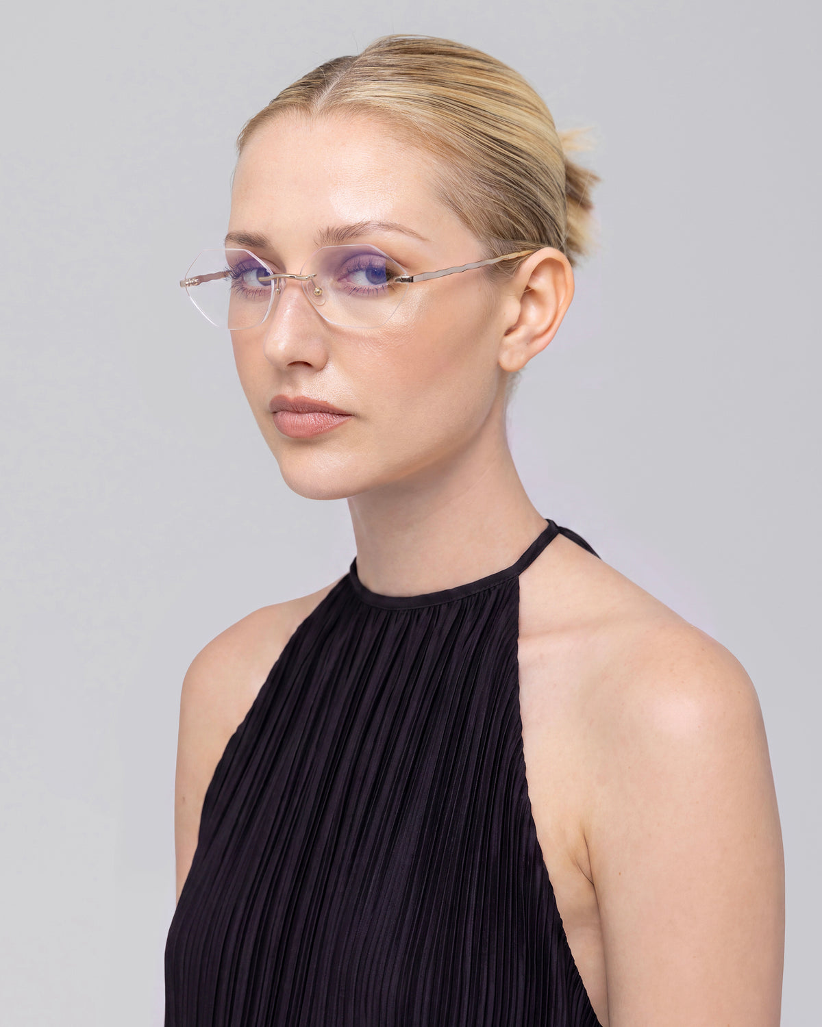A blonde woman with a sleek bun wears For Art’s Sake® Amara I rimless glasses with sculpted nylon temple tips and a sleeveless black pleated top, calmly facing the camera against a light gray background.