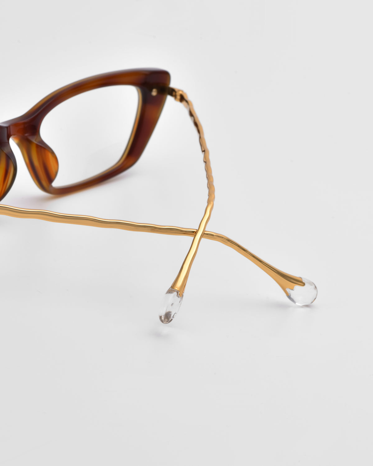 The For Art’s Sake® Amara II features a brown optical frame with gold wavy arms and sculpted nylon temple tips, shown partially resting on a white surface.