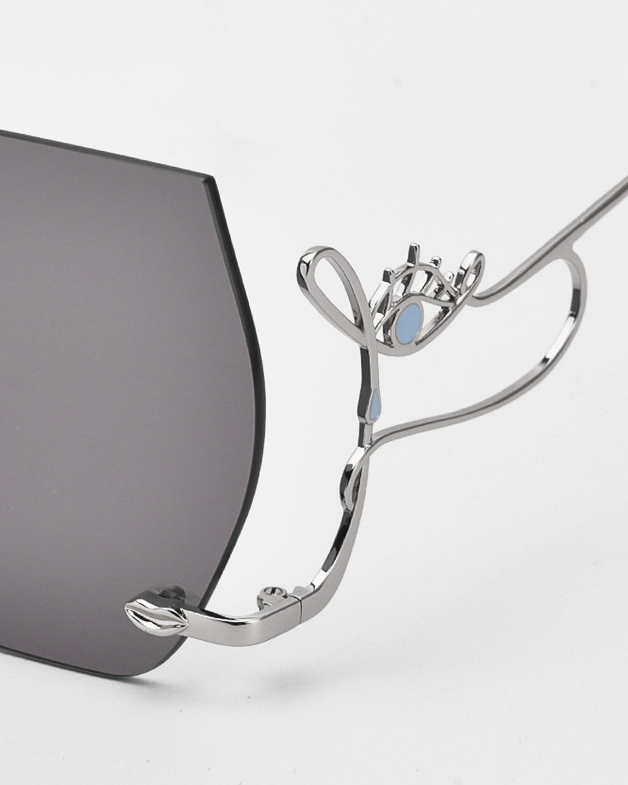 Close-up of For Art&#39;s Sake® Cry Me a River limited edition sunglasses, featuring dark lenses and an abstract silver metal eye-shaped arm with light blue enamel accents. A plain white background highlights their striking design.
