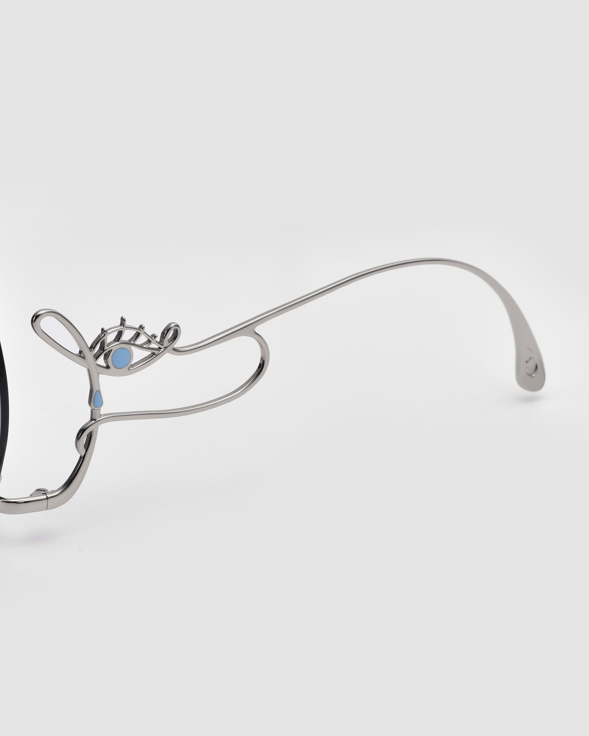 Close-up of a pair of sunglasses with a silver metal frame featuring a decorative angel wing design and small light blue accents on the upper part of the left arm, set against a light gray background.