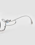 Close-up of a pair of sunglasses with a silver metal frame featuring a decorative angel wing design and small light blue accents on the upper part of the left arm, set against a light gray background.