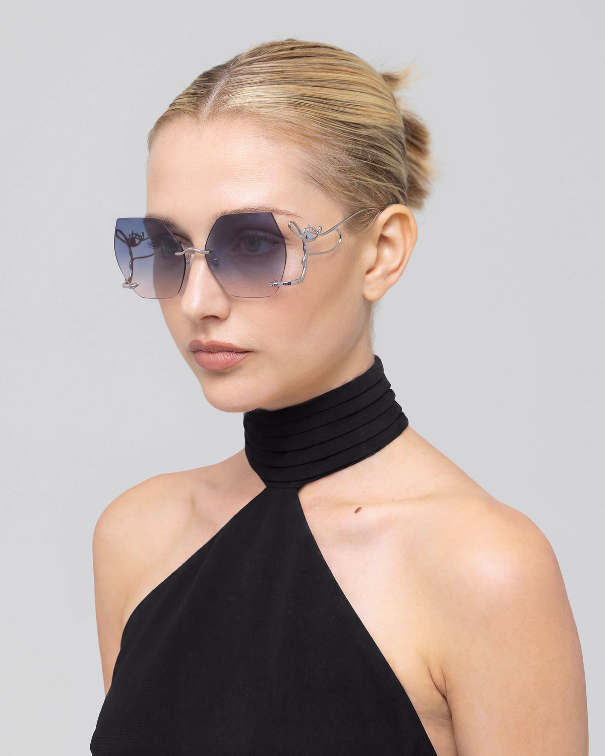 A woman with blonde hair in a bun wearing oversized square sunglasses with a blue floral pattern on the frames and a black halter top, photographed against a solid light gray background.