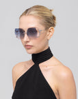 A woman with blonde hair in a bun wearing oversized square sunglasses with a blue floral pattern on the frames and a black halter top, photographed against a solid light gray background.