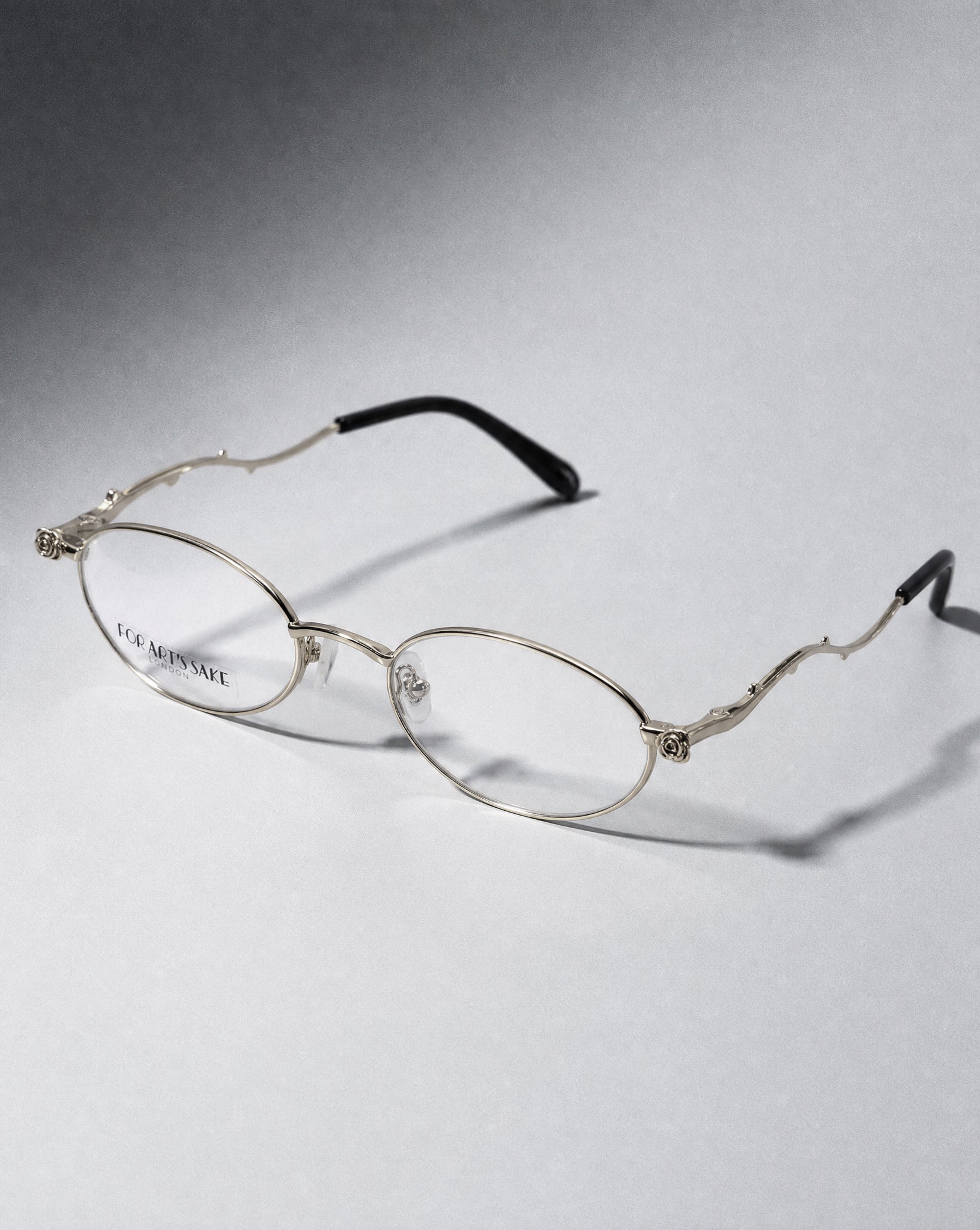 A pair of elegant, modern Thorn oval glasses by For Art's Sake® featuring thin metal frames and black temple tips rest on a light grey surface, with "FOR ARTS SAKE" printed on the left lens—true statement eyewear for any occasion.